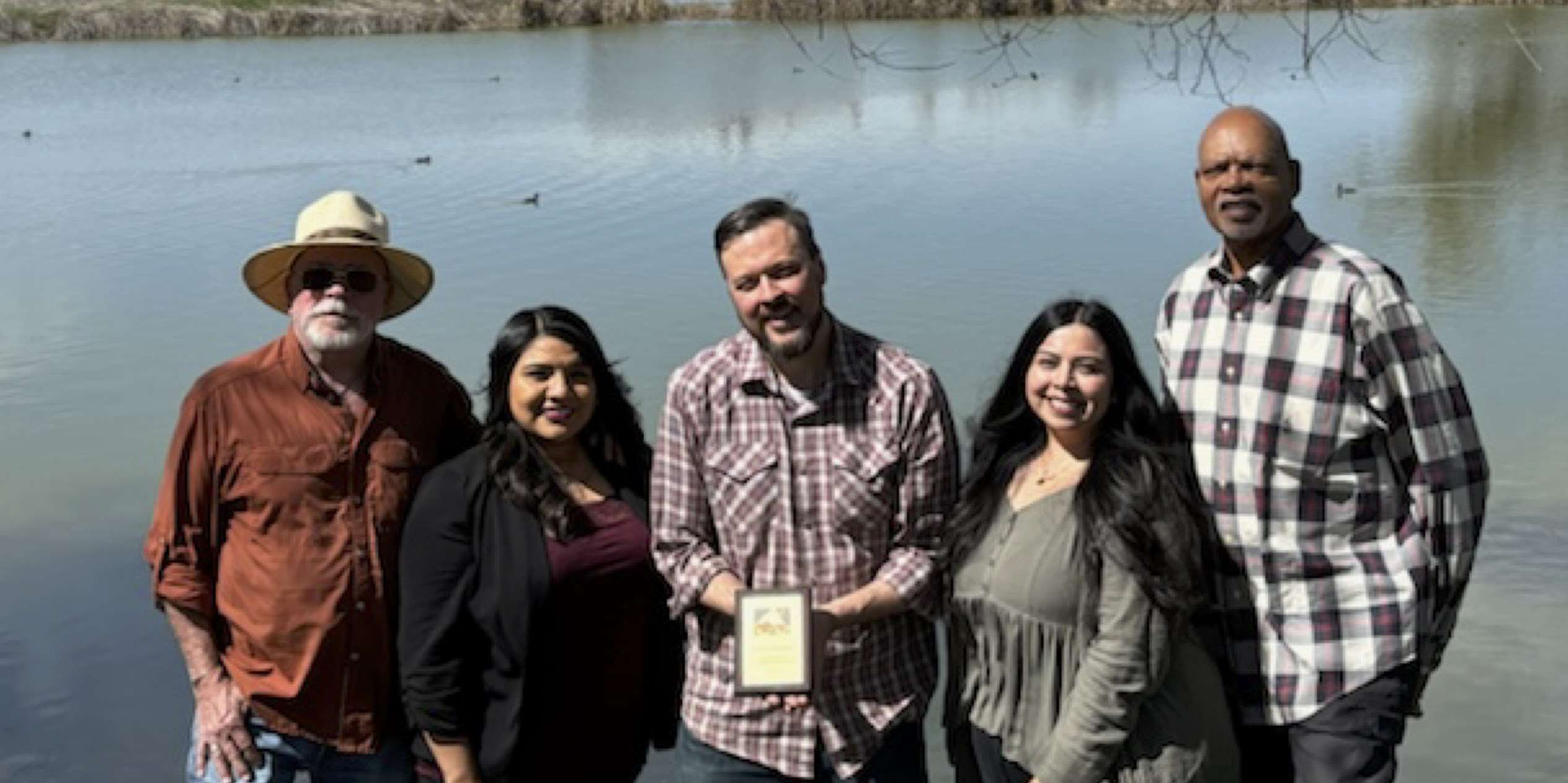 5 people stand in front of a lake smiling for the camera and holding up their plaque.