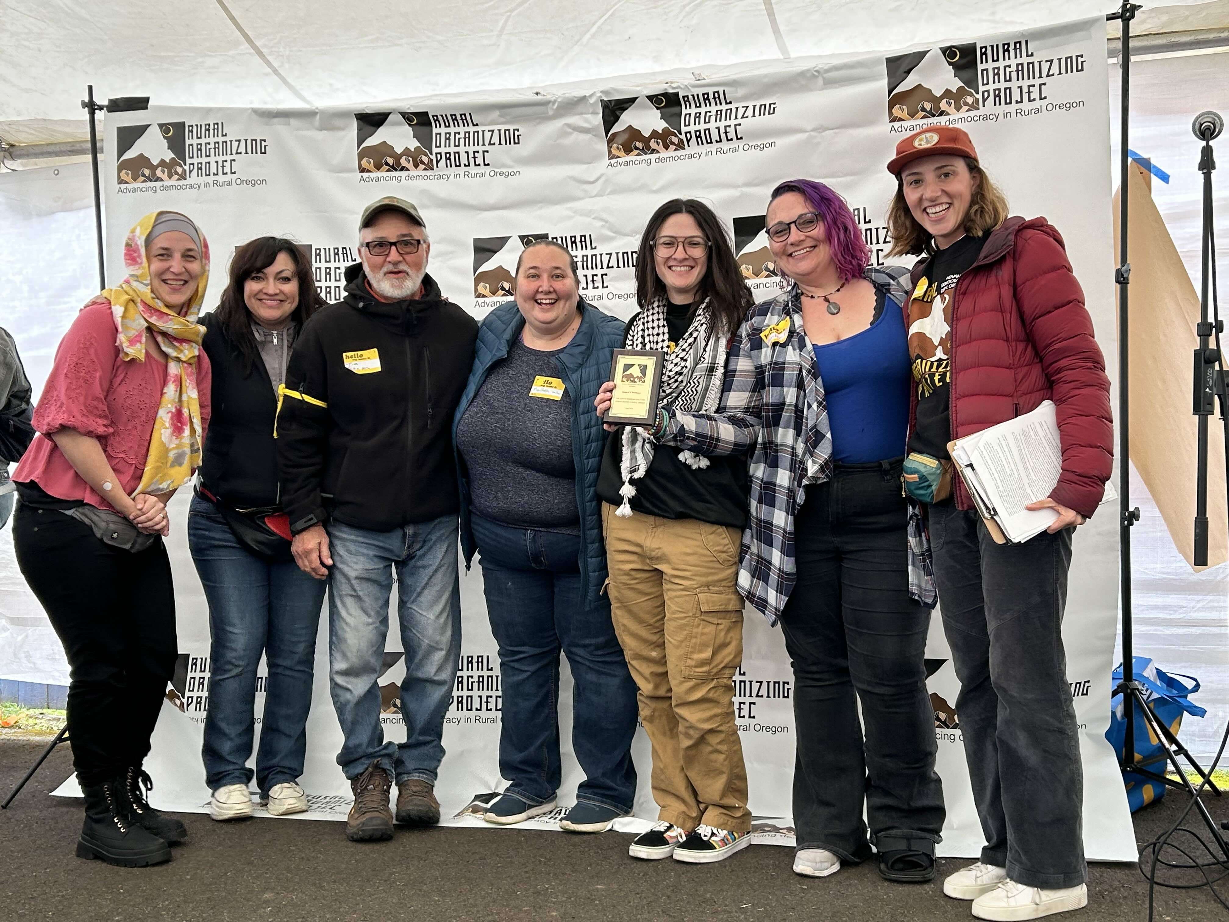 7 people pose for a photo holding their Human Dignity Award in front of an ROP banner.