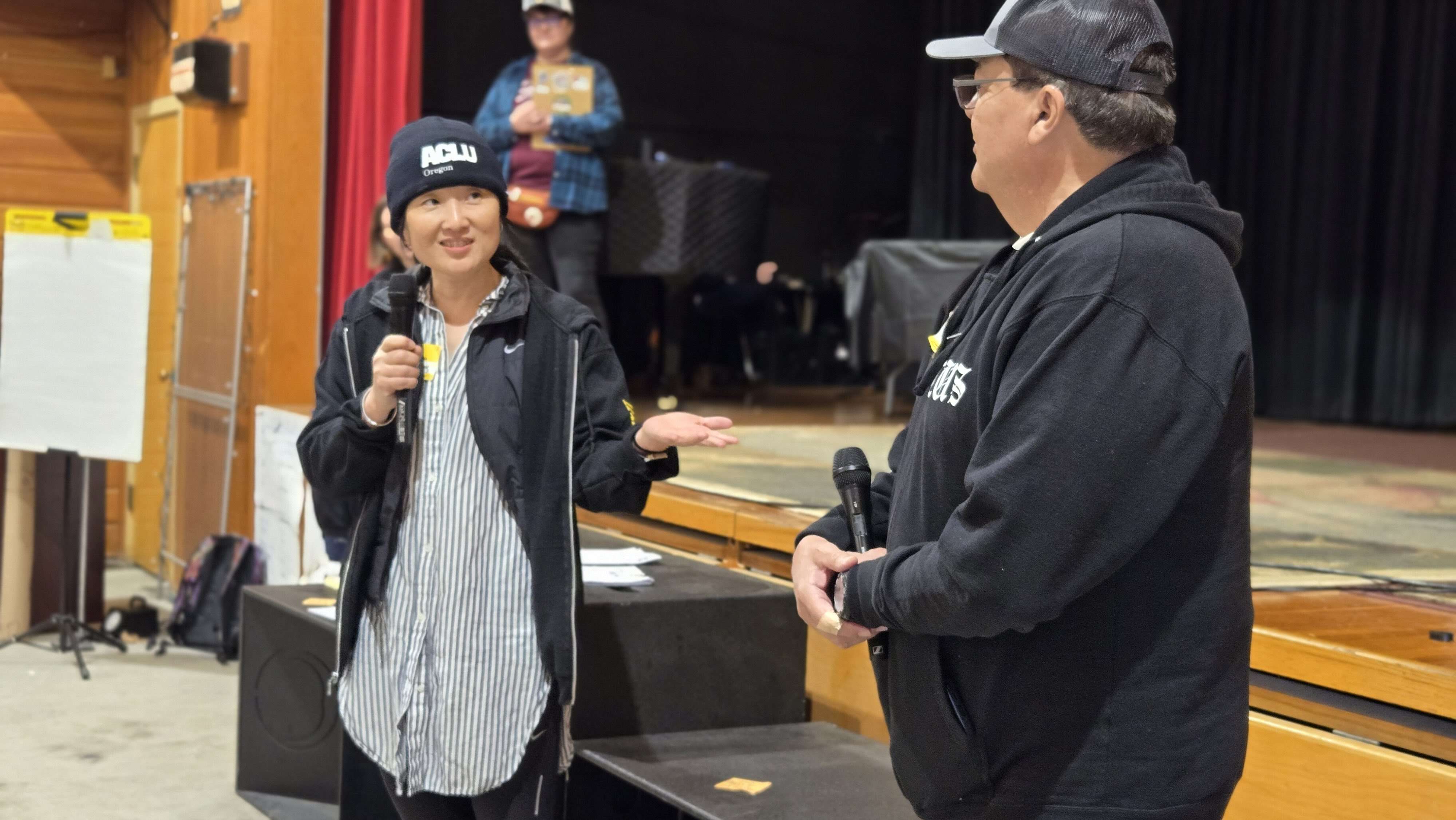 Sandy and Ramon hold microphones in front of the stage and speak to each other.